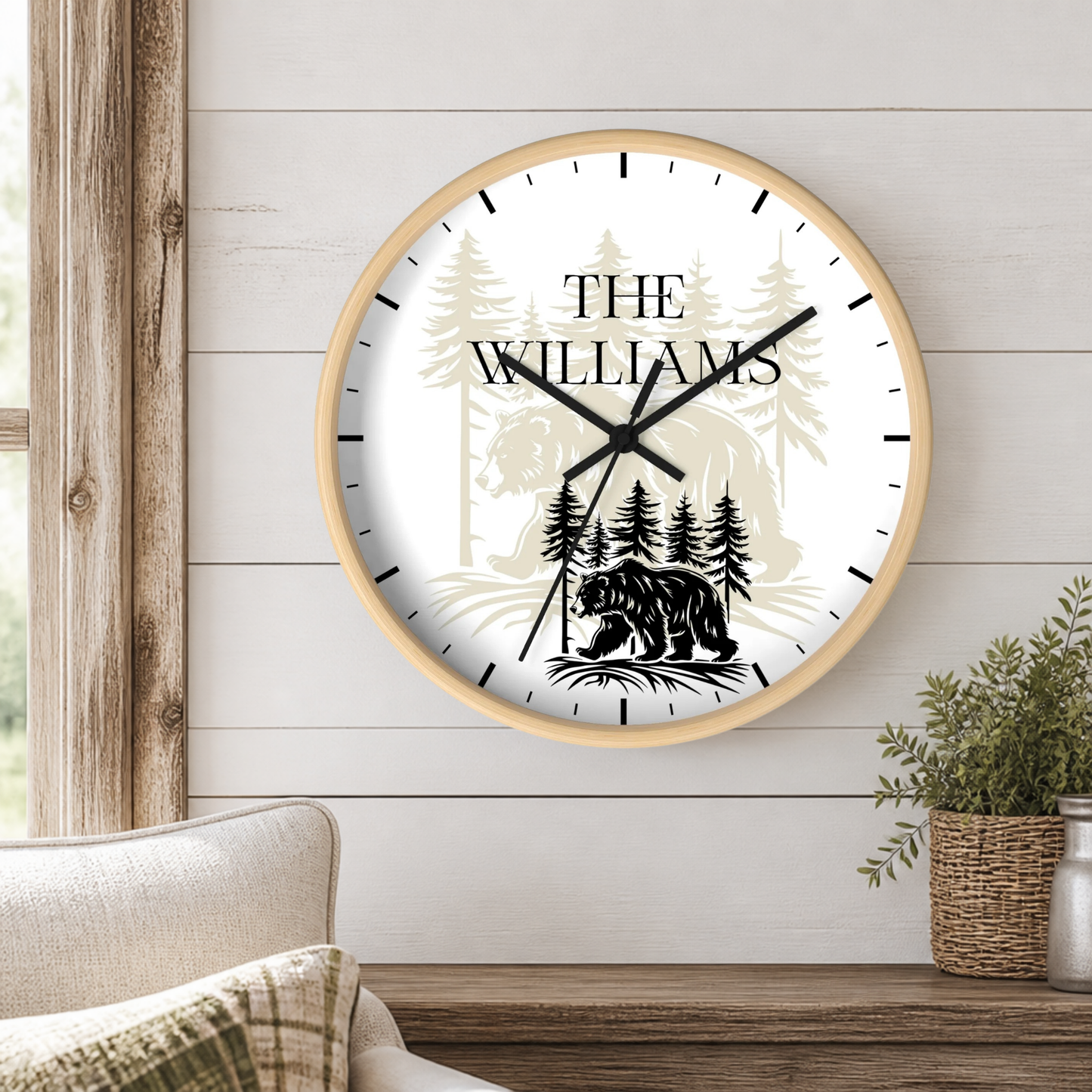 A personalized round wall clock with a light wood frame, mounted on a white shiplap wall. The clock face is white with black hands and markers, featuring a black silhouette of a bear and pine trees, and the text 'THE WILLIAMS' in a serif font.