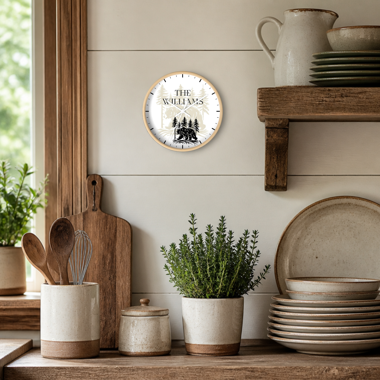 A round wall clock with a light wood frame hangs on a white shiplap wall in a rustic kitchen setting. The clock face is white with black hands and a black forest bear and tree illustration, with the text 'THE WILLIAMS' at the top.