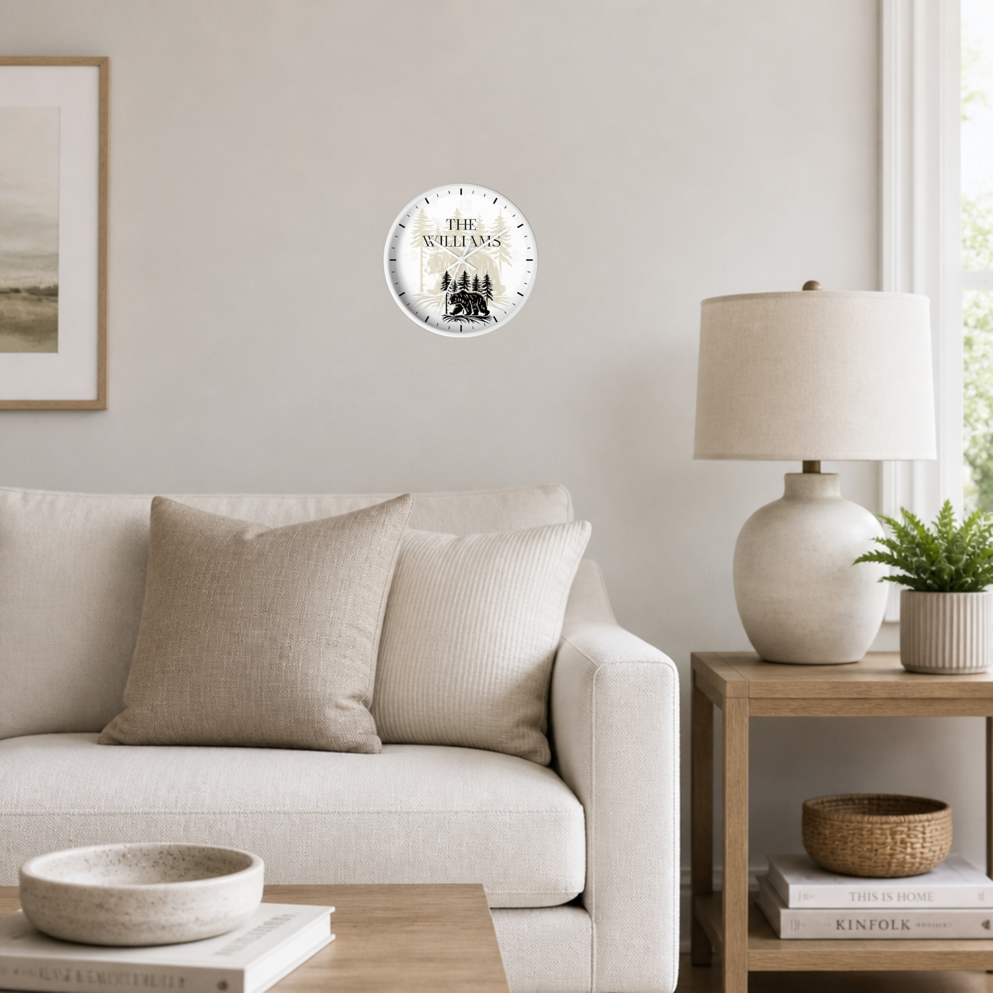 A round wall clock with a distressed white face and black text reading 'THE Williams' is mounted on a light beige wall in a styled living room setting with a sofa, side table, and lamp.