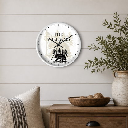 A personalized round wall clock with a white face, black hands, and a black bear silhouette over a beige forest pattern. The clock is mounted on a light beige shiplap wall above a wooden side table with a plant in a ceramic pot.