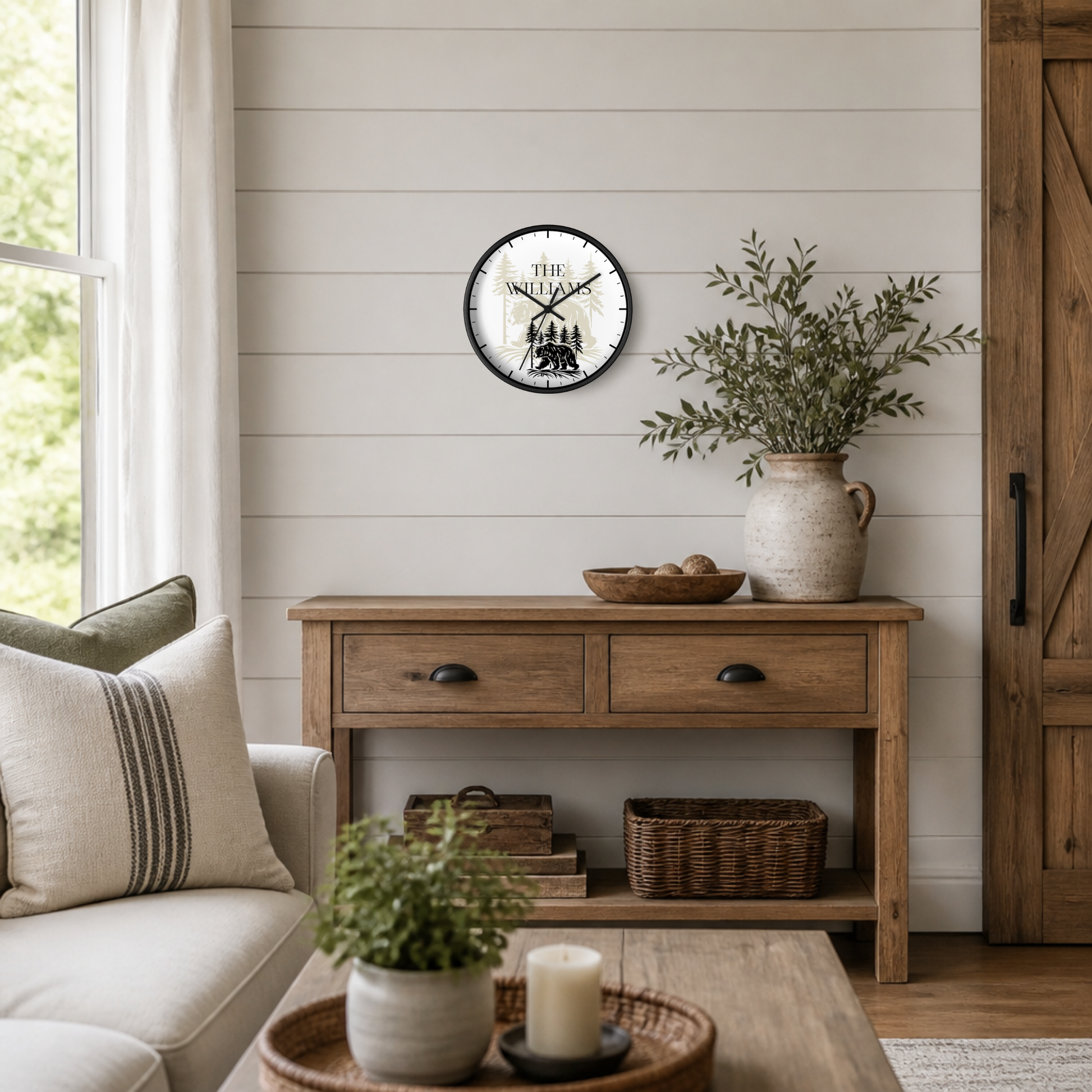 A round wall clock with a black frame and white face featuring the text 'THE Williams' and a black silhouette illustration, mounted on a white shiplap wall above a rustic wooden console table in a styled living room.