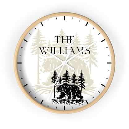 A personalized round wall clock with a light natural wood frame, featuring a white face with black hands and hour markers. The clock face is printed with a black bear silhouette, pine trees, and the text 'THE WILLIAMS' in a serif font.