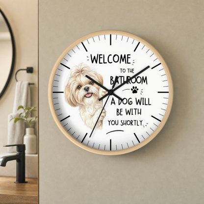 A round wall clock with a light wood frame and a white face, mounted on a beige wall in a bathroom setting. The clock features a drawing of a fluffy white dog and the text 'WELCOME TO THE BATHROOM A DOG WILL BE WITH YOU SHORTLY' with black hour markers and hands.