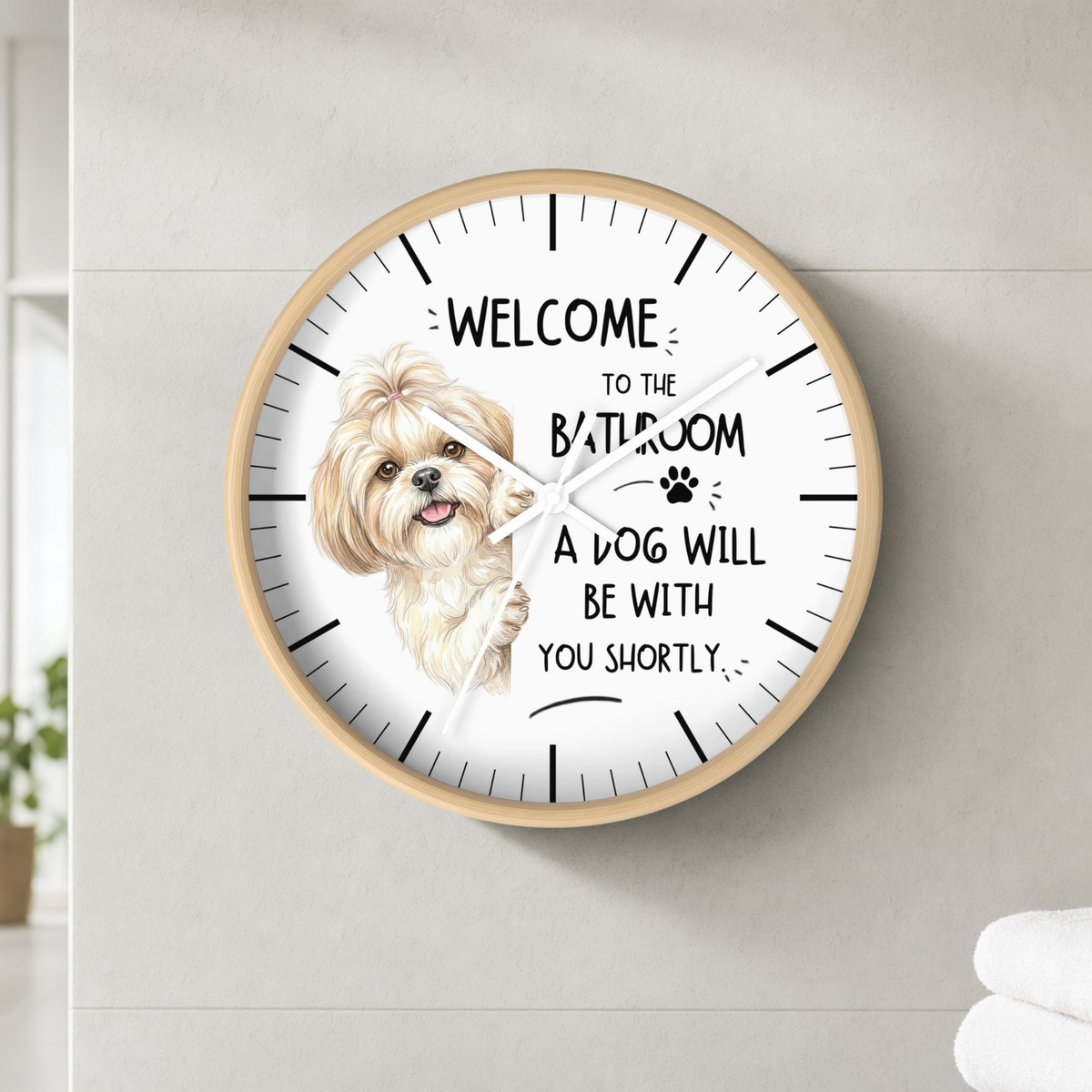 A round wall clock with a light wood frame and a white face, featuring a drawing of a small fluffy dog and the text 'WELCOME TO THE BATHROOM A DOG WILL BE WITH YOU SHORTLY'. The clock is mounted on a light gray tiled wall.