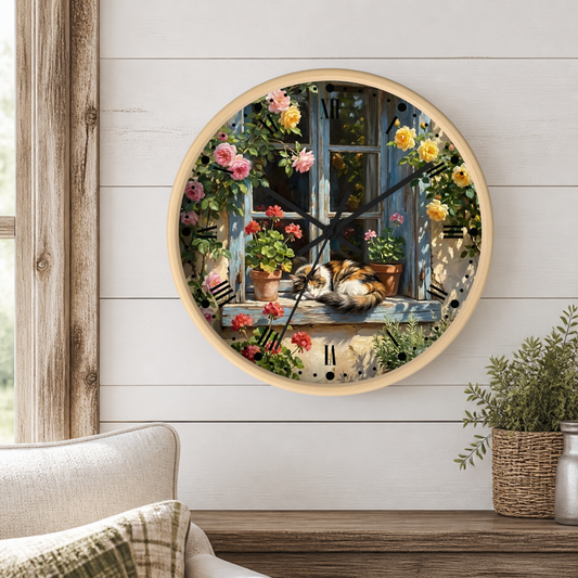 A round wall clock with a light wood frame hangs on a white shiplap wall above a wooden table. The clock face features a detailed illustration of a cozy cat sleeping on a window sill surrounded by blooming pink, red, and yellow flowers.