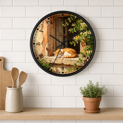 A round wall clock with a black frame hangs on a white subway-tile kitchen wall above a wooden shelf. The clock face features a scenic print of an orange cat sleeping by a rustic window with green vines.