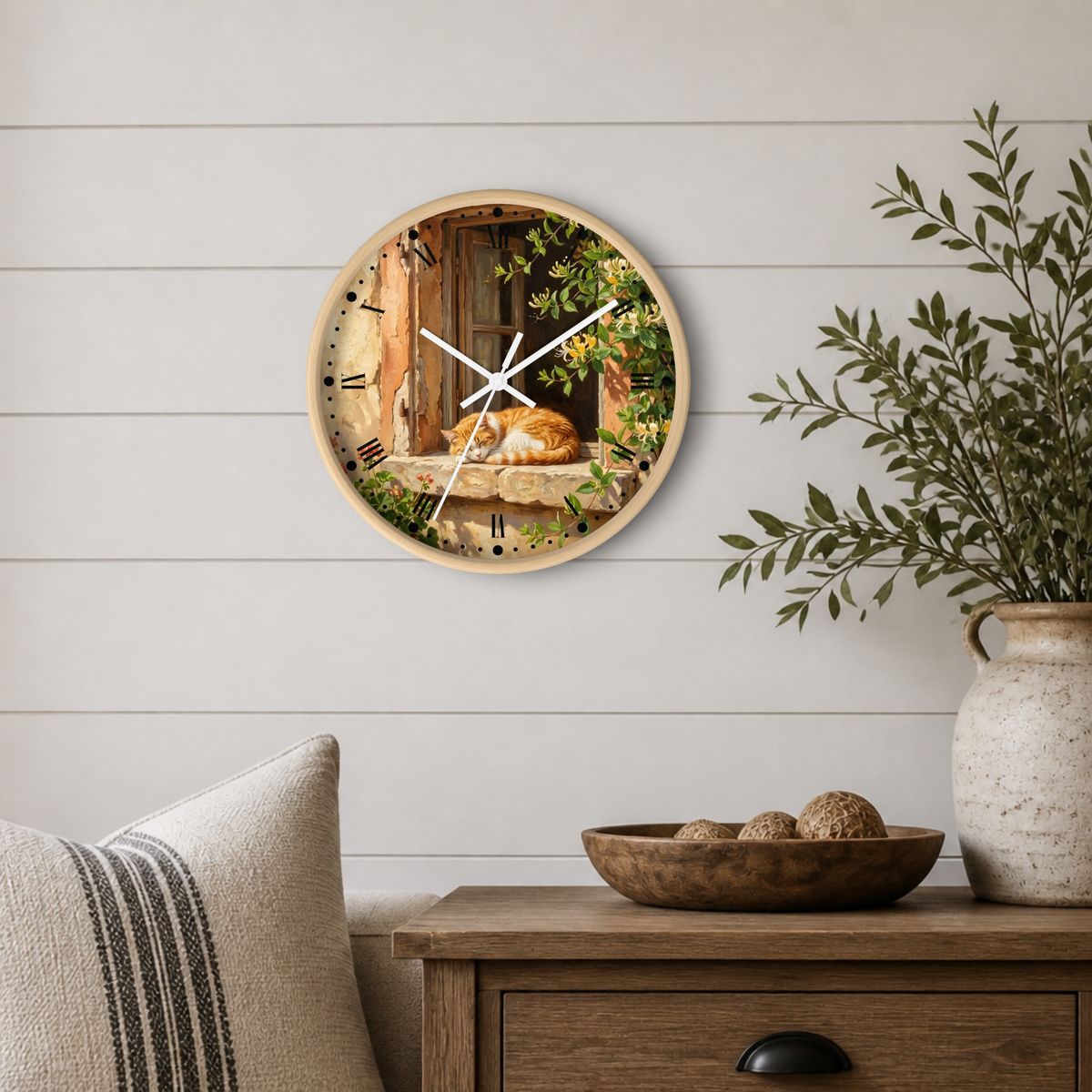 Round wall clock with a natural wood frame and a printed scene of a cat resting on a windowsill, styled on a white shiplap wall above a wooden side table with a plant and decorative bowl.