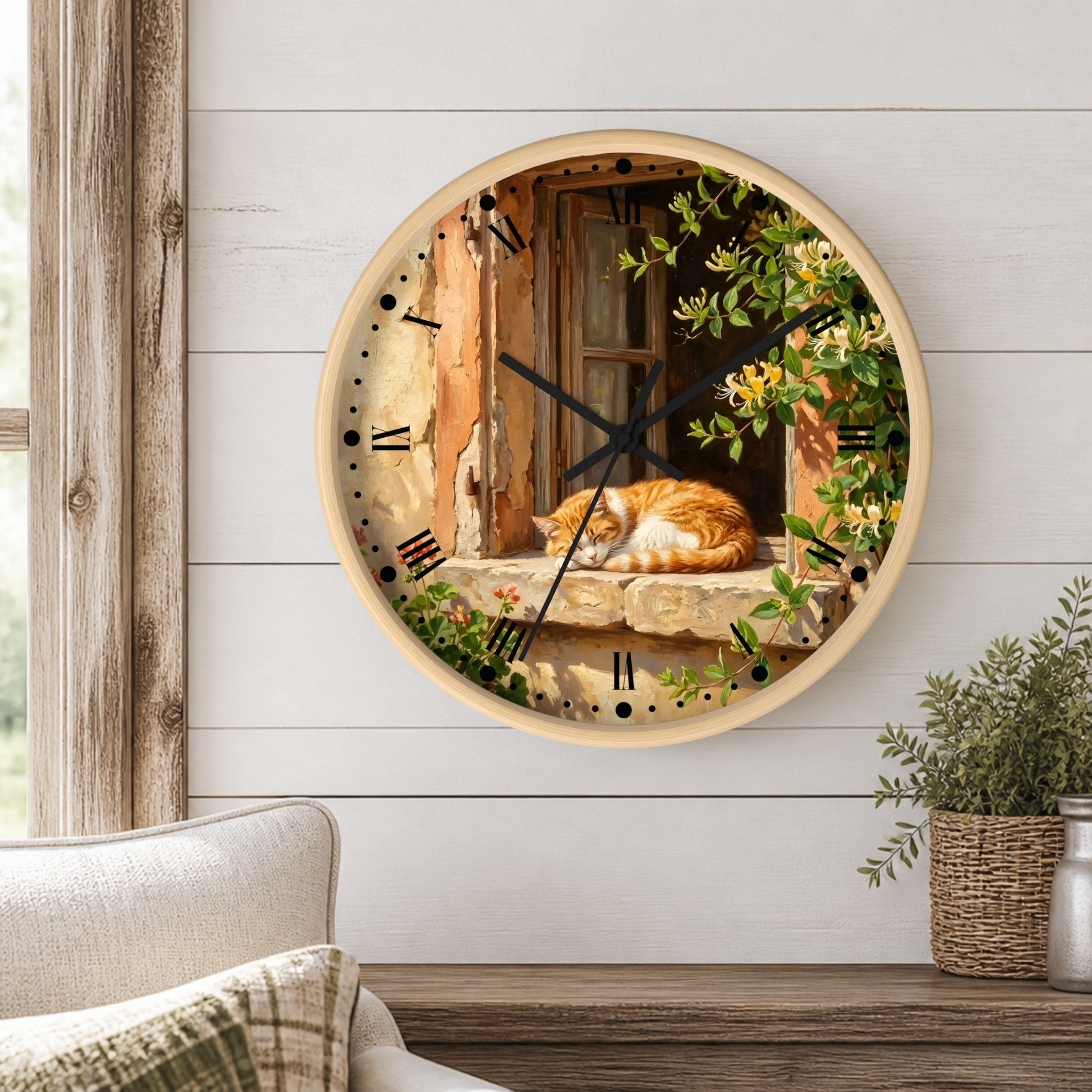 Round wall clock with a light wood frame, featuring a printed design of an orange cat sleeping by a rustic window with green foliage. The clock is mounted on a white wood-paneled wall above a wooden shelf with a potted plant.