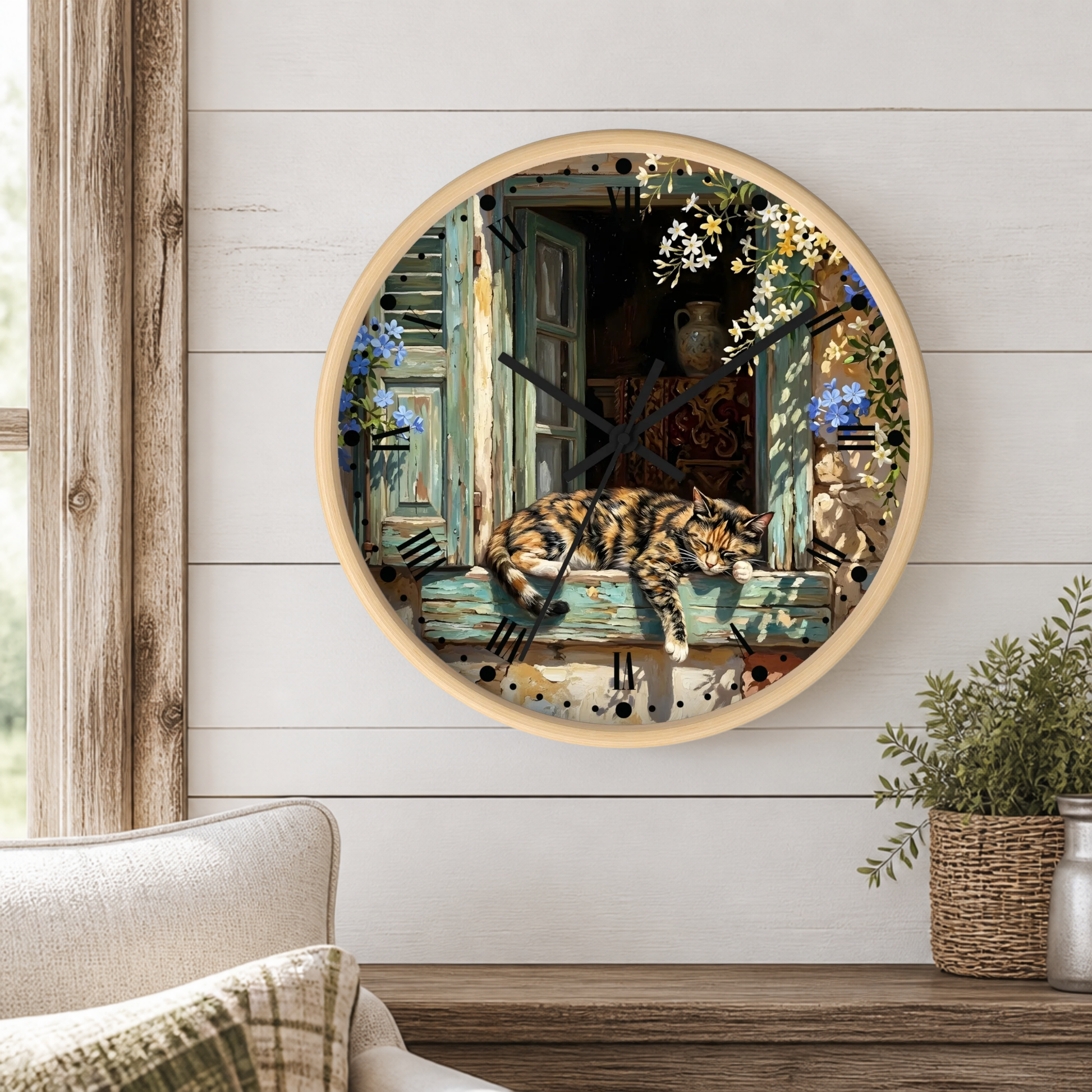 Round wall clock with a light wood frame, featuring a printed illustration of a tortoiseshell cat resting on a weathered blue-green wooden window sill with flowers and a bird perched nearby. The clock is mounted on a white shiplap wall above a wooden mantel with a potted plant.
