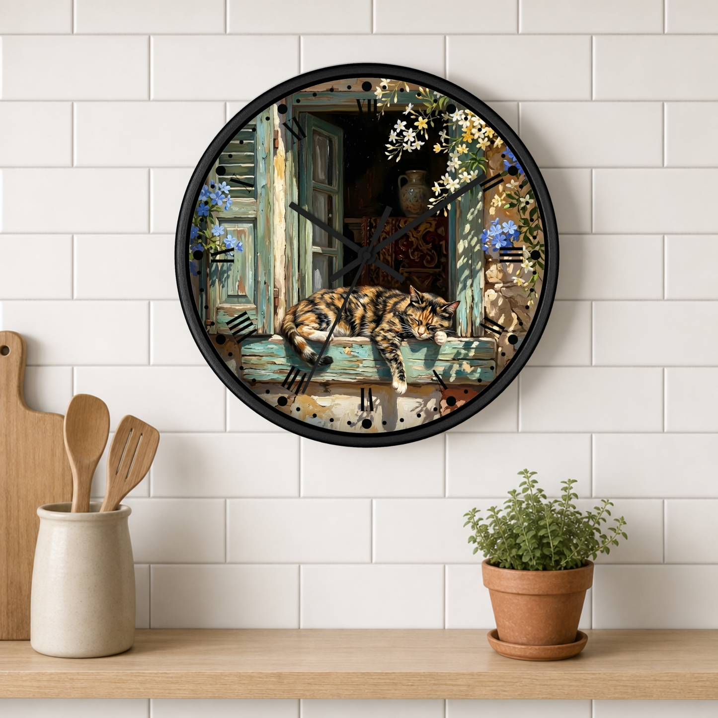 A round wall clock with a black frame and a detailed illustration of a torte cat sleeping on a weathered window sill, mounted on a white subway tile wall above a wooden shelf with kitchen utensils and a potted plant.