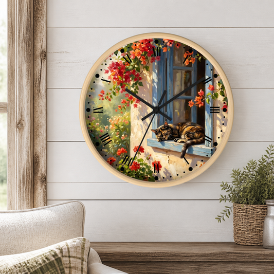 A round wall clock with a light wood frame hangs on a white shiplap wall above a wooden table. The clock face features a scenic print of a cat resting on a blue window sill surrounded by red flowers, with black clock hands and Roman numerals.