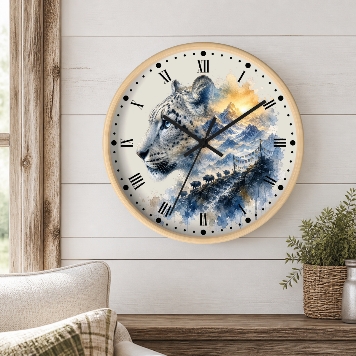A round wall clock with a light wood frame hangs on a white shiplap wall in a styled room. The clock face features a watercolor illustration of a snow leopard over blue mountains and a river, with black Roman numerals and hands.