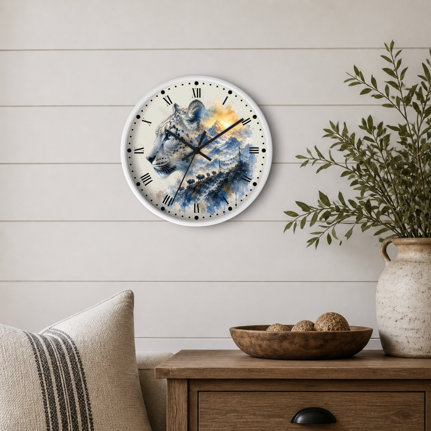 A round wall clock with a painted snow leopard design in blue, white, and gold tones, mounted on a light beige shiplap wall above a wooden side table with a plant and decorative bowl.