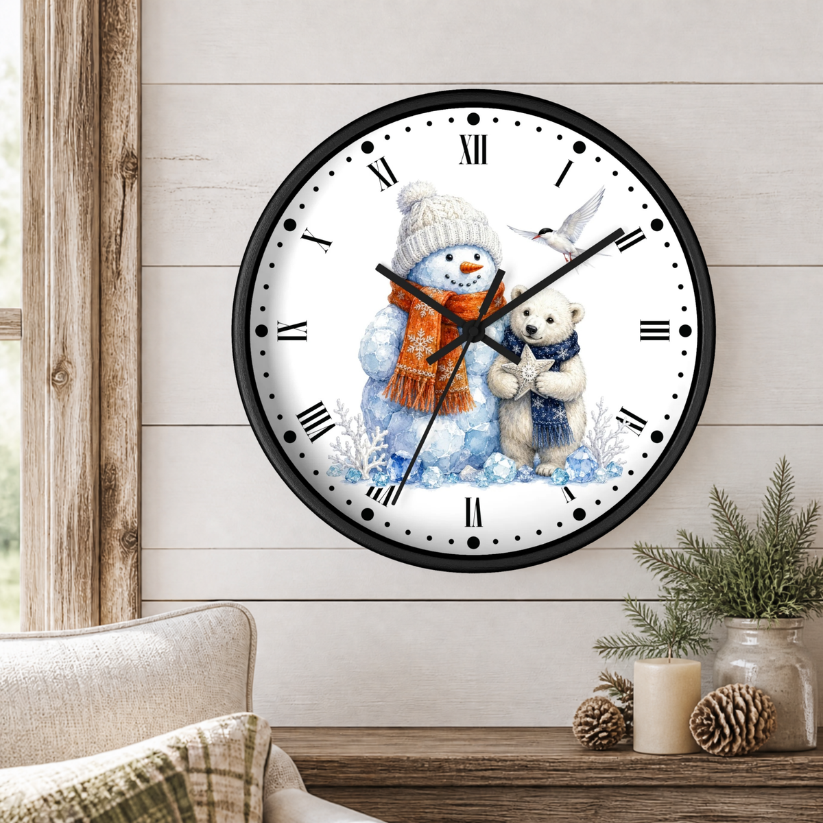 A round wall clock with a black frame and a white face, featuring a watercolor illustration of a snowman in a beige knit hat and orange scarf hugging a small white bear. The clock is mounted on a light wood-paneled wall above a wooden table with a candle, pinecones, and a small plant.