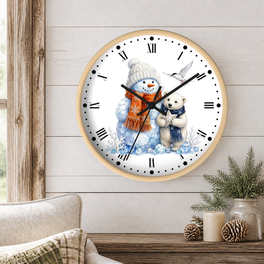 A round wall clock with a light wood frame hangs on a white shiplap wall above a wooden table. The clock face features a winter illustration of a snowman in a beige knit hat and orange scarf hugging a white bear, with a white bird at the top right.