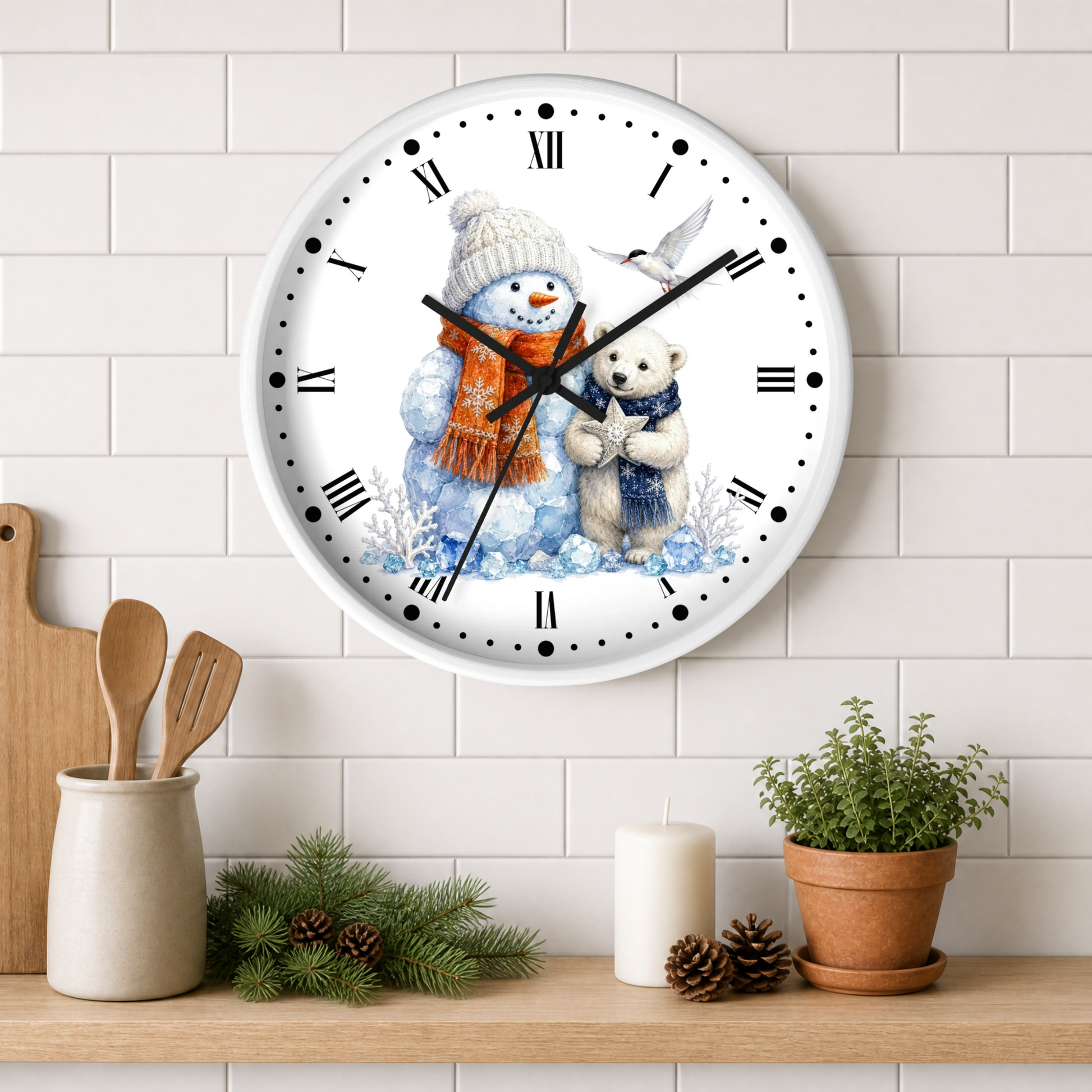 A round wall clock with a white face and black Roman numerals, featuring a winter illustration of a snowman in an orange scarf hugging a polar bear, displayed on a white subway tile wall above a shelf with kitchen props.