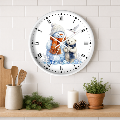 A round wall clock with a white face and black Roman numerals, featuring an illustration of a snowman in a beige hat and orange scarf hugging a small white bear. The clock is mounted on a white subway tile wall above a wooden shelf styled with kitchen utensils, pine branches, a candle, and a potted plant.