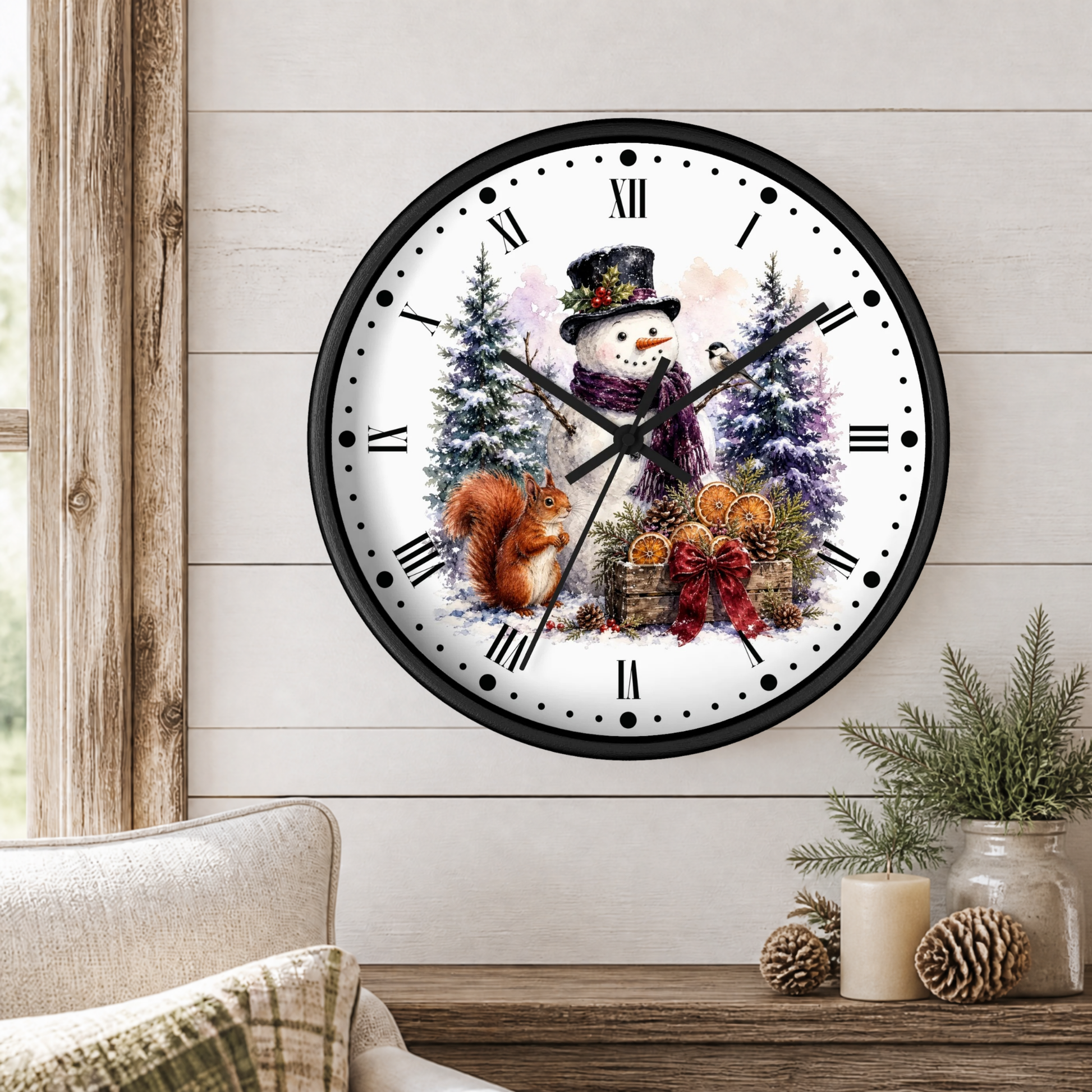 A round wall clock with a winter holiday illustration, featuring a snowman, squirrel, pine trees, and ornaments on a white face with black Roman numerals, shown hanging on a light wood paneled wall above a rustic table with candles and greenery.