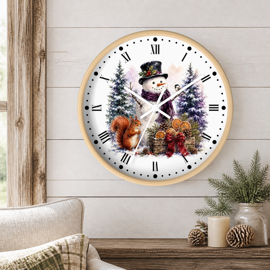 A round wall clock with a light wood frame and a winter holiday illustration featuring a snowman, squirrel, pine trees, and ornaments, mounted on a white wood-paneled wall above a rustic table with candles and greenery.