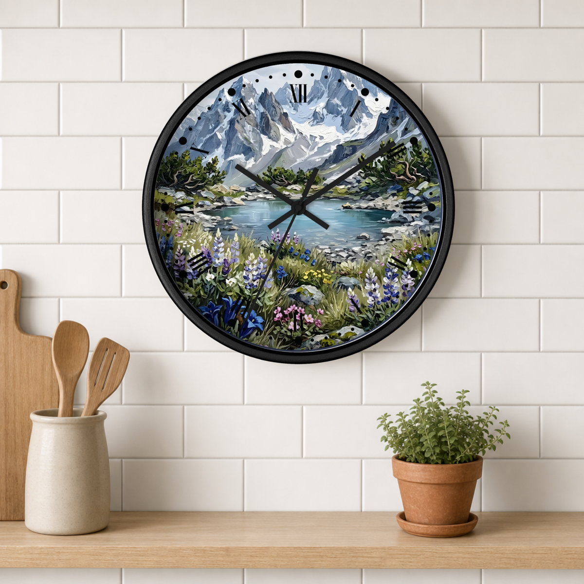 A round wall clock with a black frame hangs on a white subway-tile kitchen wall above a wooden shelf. The clock face features a colorful scenic print of a mountain lake surrounded by wildflowers.