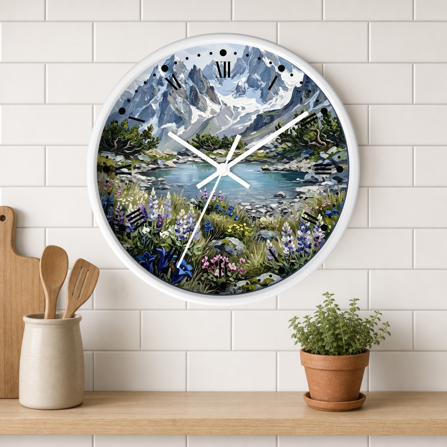 A round wall clock with a white frame and a scenic landscape print of mountains, a lake, and wildflowers, mounted on a white subway tile wall above a wooden shelf with kitchen utensils and a potted plant.