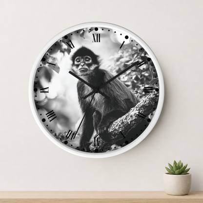 A round wall clock with a white frame and a black-and-white photographic print of a monkey sitting on a tree branch, mounted on a light beige wall above a wooden surface with a small potted succulent.
