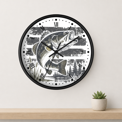 A round wall clock with a black frame and a white face featuring a detailed black-and-gray illustration of a largemouth bass over a striped background with trees and reeds. The clock is mounted on a light beige wall above a light wood shelf, with a small potted succulent on the right.