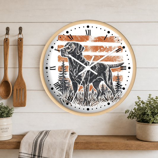 A round wall clock with a light wood frame hangs on a white shiplap wall above a wooden shelf. The clock face features a black Labrador retriever standing in front of distressed orange and white horizontal stripes, with black Roman numerals and clock hands.