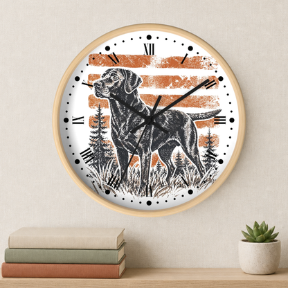 A round wall clock with a light wood frame hangs on a beige wall above a shelf with stacked books and a small potted succulent. The clock face features a black-and-white illustration of a dog standing in grass with pine trees, set against a white background with distressed orange horizontal stripes and black Roman numerals.