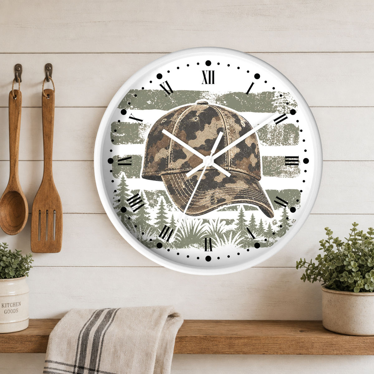 A round wall clock with a white frame and a printed design of a camouflage baseball cap over a forest and distressed green-and-white stripe background. The clock hangs on a white wood plank wall above a wooden shelf with kitchen utensils, plants, and a towel.