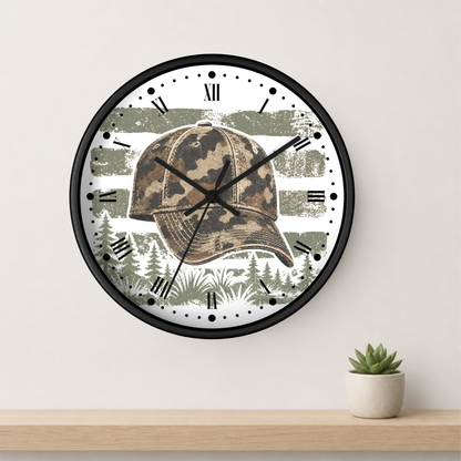 A round wall clock with a black frame and a printed design featuring a camouflage baseball cap over a forest scene with horizontal olive-green stripes. The clock is mounted on a light beige wall above a wooden shelf with a small potted succulent.