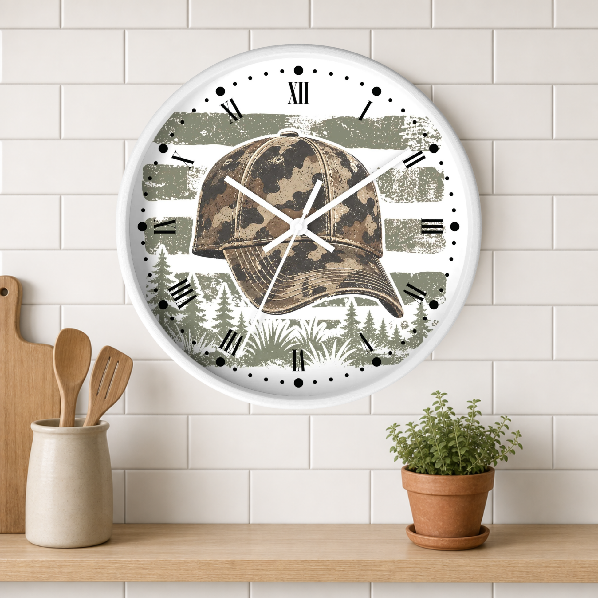 A round wall clock with a white frame hangs on a white subway tile wall above a wooden shelf. The clock face features a camouflage baseball cap over distressed olive green and gray brushstroke graphics with a pine tree pattern at the bottom.