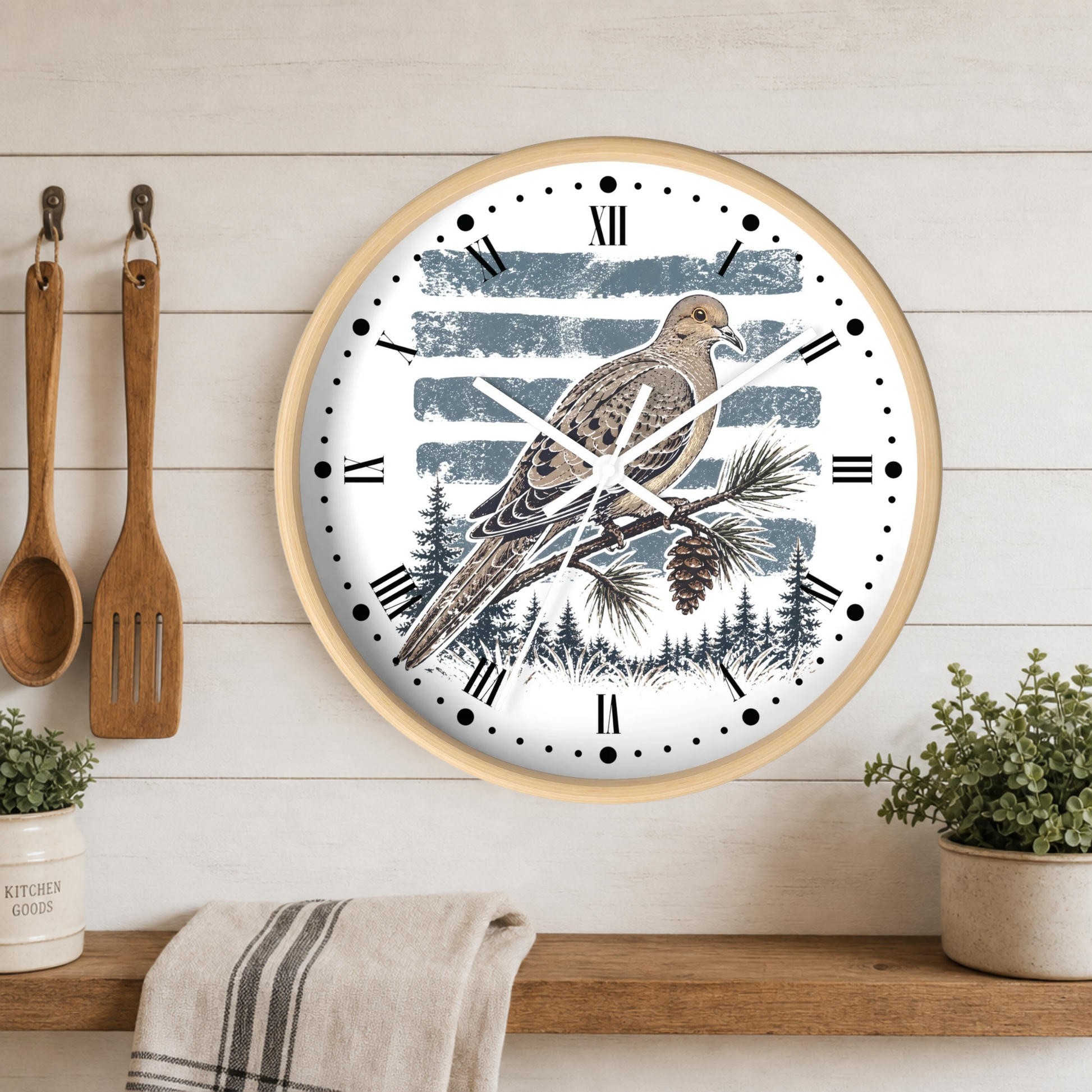 A round wall clock with a light wood frame hangs on a white shiplap wall above a wooden shelf. The clock face features a detailed illustration of a dove perched on a pine branch with a pinecone, set against a background of blue-gray horizontal brushstrokes and a forest silhouette.