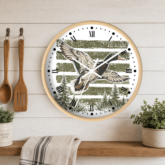 Round wall clock with a light wood frame, featuring a rustic hunting scene with a mallard duck flying over green pine trees and horizontal stripes. The clock is mounted on a white wood-paneled wall above a wooden shelf with kitchen utensils, plants, and a towel.