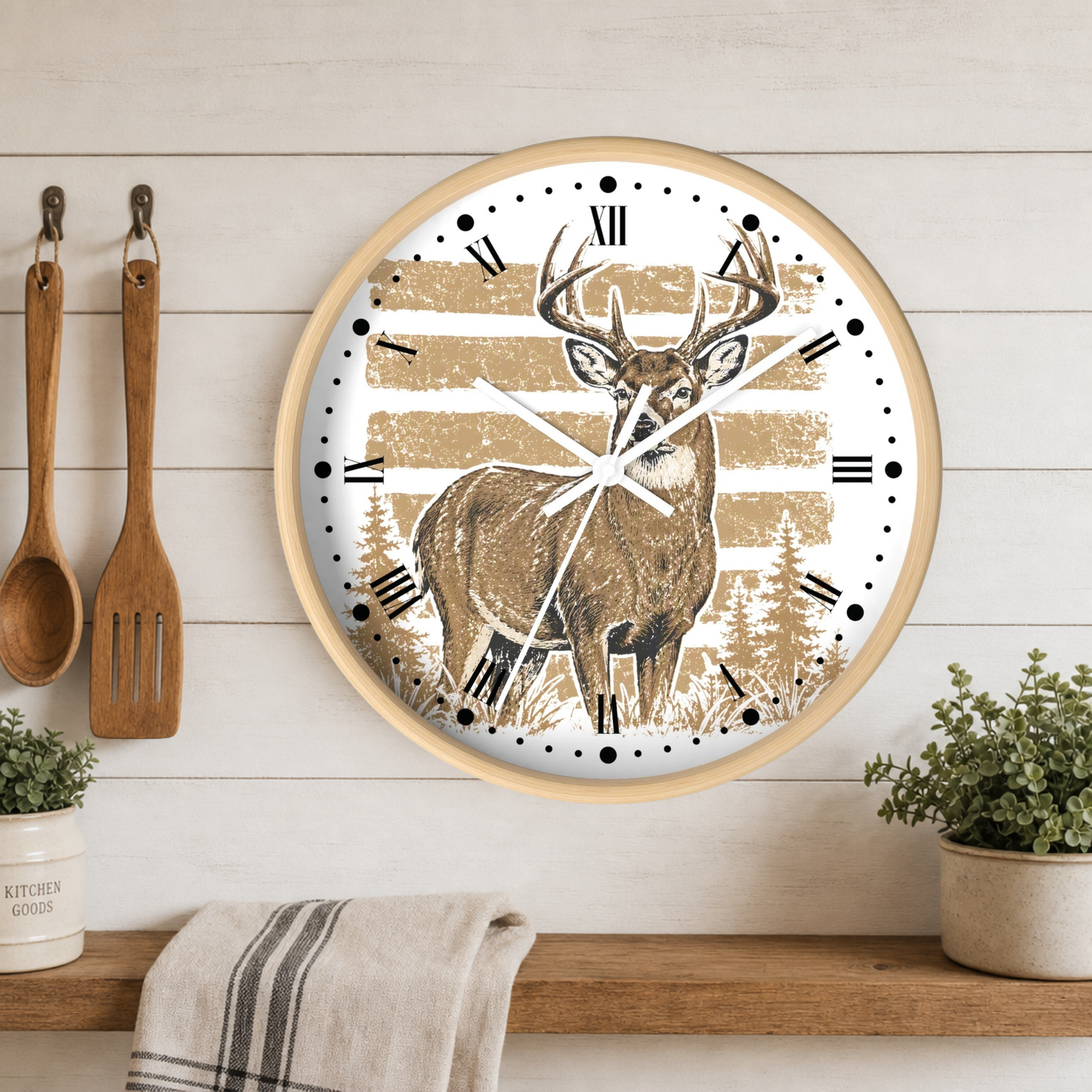 Round wall clock with a light wood frame, featuring a deer illustration over tan and white horizontal stripes with black Roman numerals. The clock is mounted on a white wood-paneled wall above a wooden shelf with kitchen utensils, plants, and a towel.