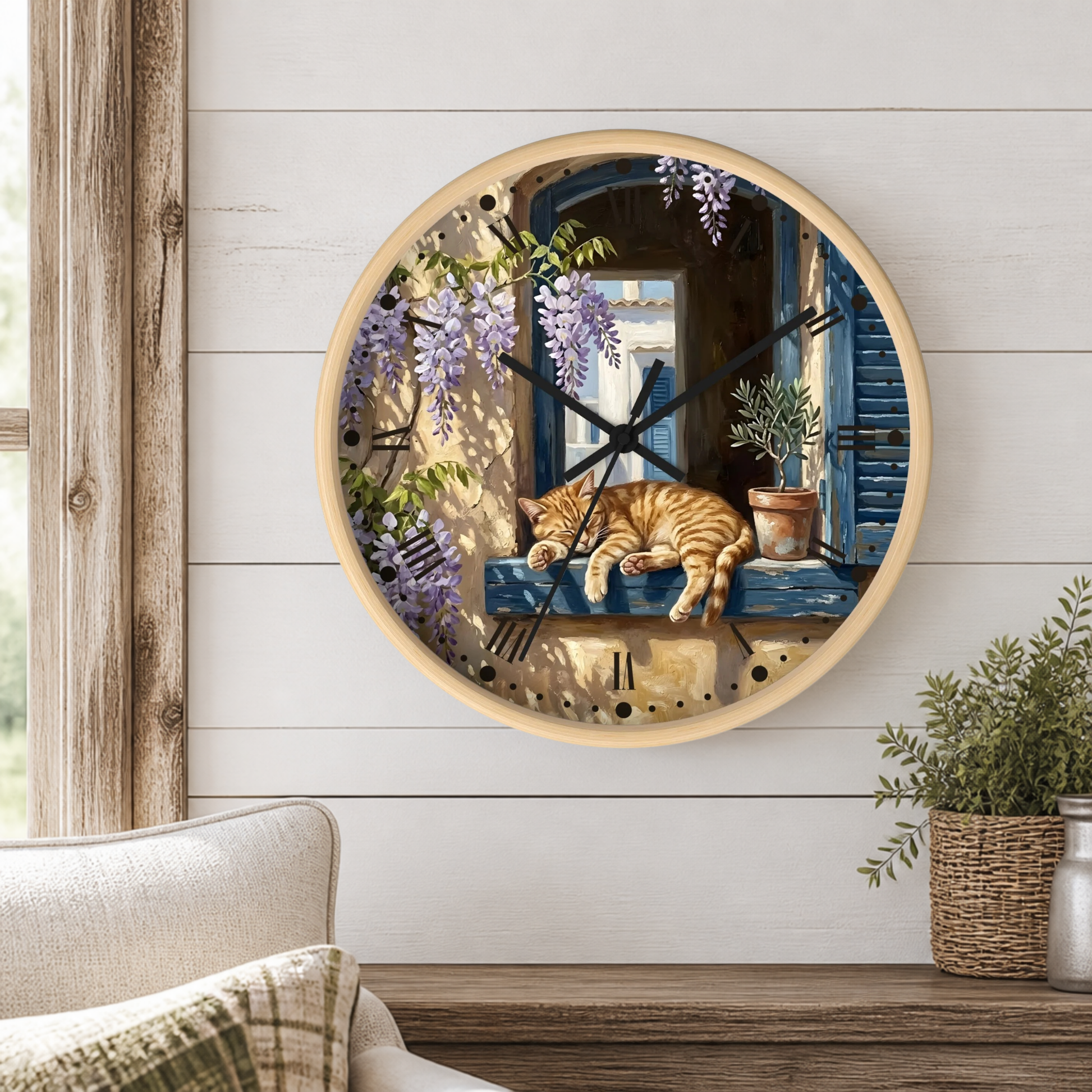 A round wall clock with a light wood frame hangs on a white shiplap wall above a wooden table. The clock face features a detailed illustration of an orange tabby cat sleeping on a blue windowsill, surrounded by purple wisteria flowers and potted plants.