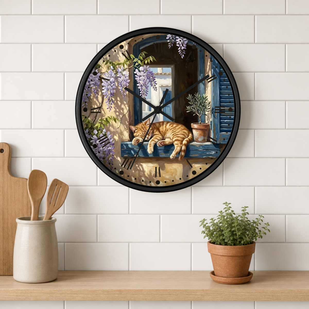 A round wall clock with a black frame hangs on a white subway-tile kitchen wall above a wooden shelf. The clock face features a detailed illustration of an orange tabby cat sleeping on a blue windowsill with purple wisteria flowers.