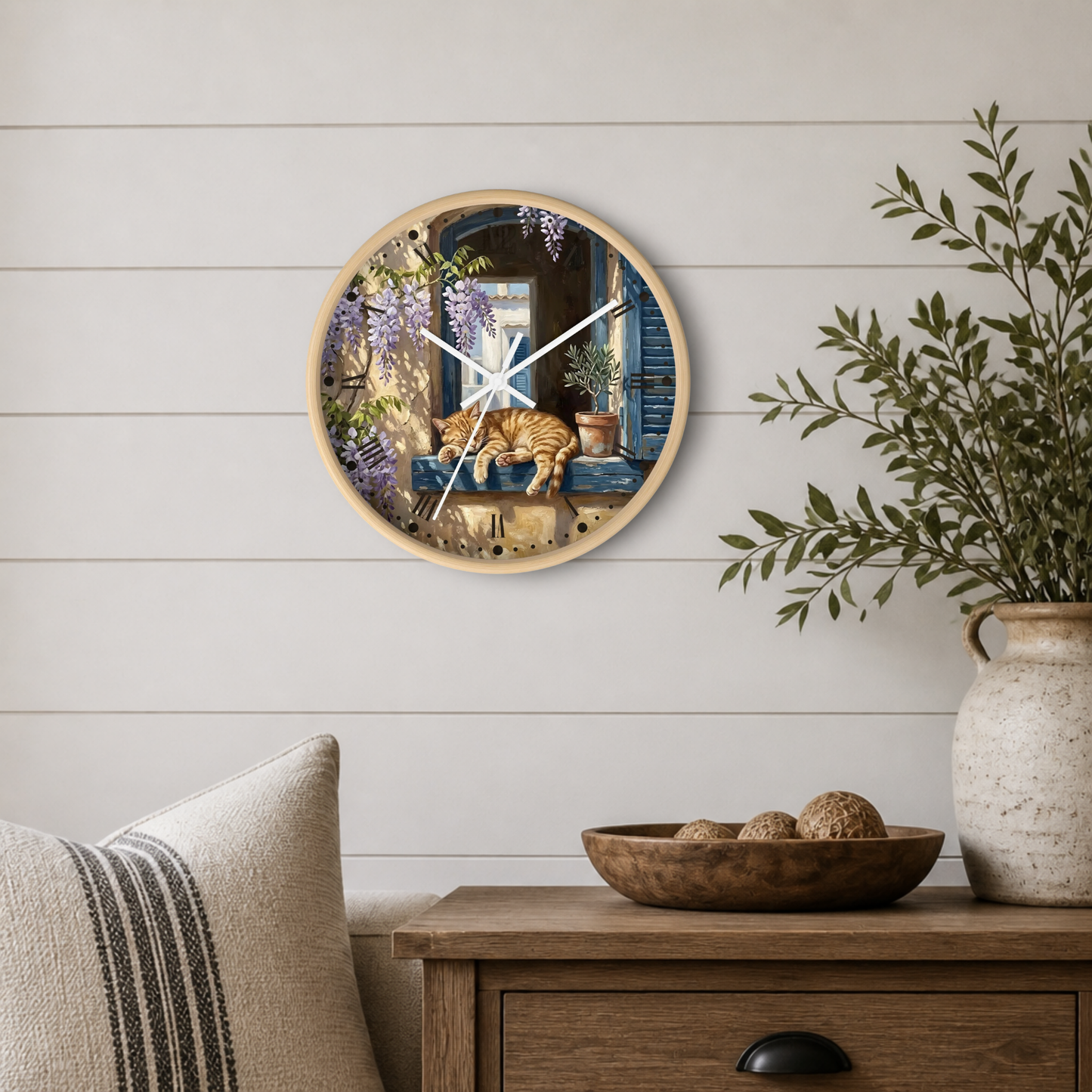 A round wall clock with a light wood frame hangs on a white shiplap wall above a wooden side table. The clock face features a detailed illustration of a cat lounging on a windowsill with wisteria and a blue door in the background.