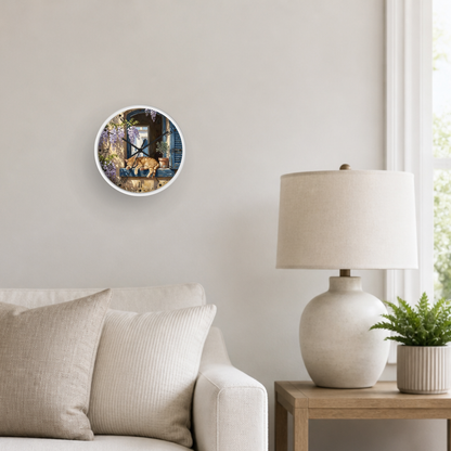 A round wall clock with a white frame hangs on a light beige wall in a styled living room setting, next to a white table lamp on a wooden side table and a small potted green plant.