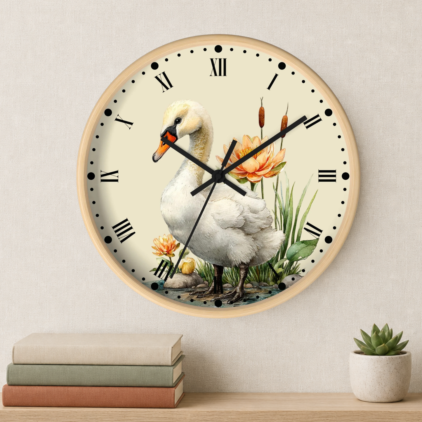 A round wall clock with a light wood frame and a cream dial, featuring a printed illustration of a white swan standing among green reeds and orange flowers. The clock is mounted on a beige wall above a shelf with stacked books and a small potted succulent.