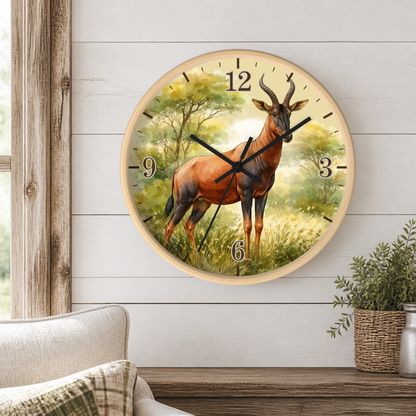 A round wall clock featuring a painted red duiker antelope in a grassy woodland scene, mounted on a white shiplap wall above a wooden console table with a potted plant and part of a sofa visible.