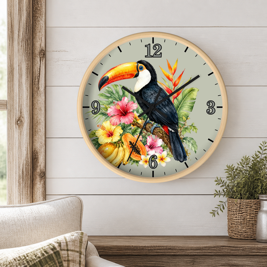 A round wall clock with a light wood frame hangs on a white shiplap wall above a wooden table. The clock face features a detailed illustration of a toucan perched among tropical flowers and leaves on a pale gray background.