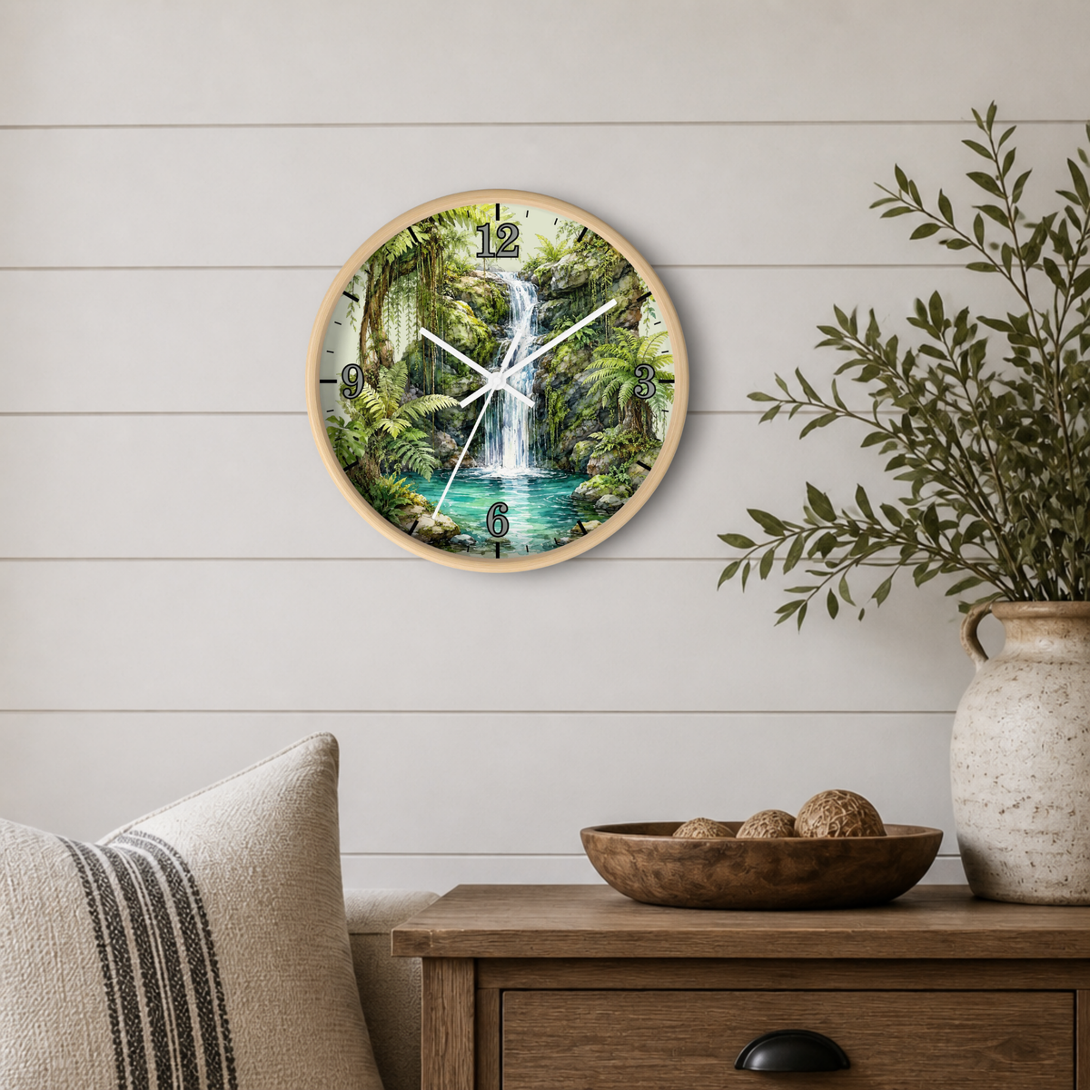 A round wall clock with a natural wood frame and a tropical waterfall landscape print, mounted on a white shiplap wall above a wooden side table with a vase and pillows.