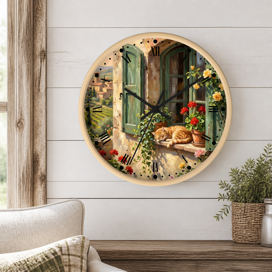 A round wall clock with a light wood frame hangs on a white shiplap wall above a wooden table. The clock face features a scenic painting of a Tuscan window with green shutters, red flowers, and a sleeping orange cat.