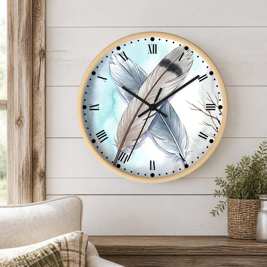 A round wall clock with a light wood frame hangs on a white shiplap wall above a wooden table. The clock face features a watercolor design of two feathers in soft blue, gray, and white tones, with black Roman numerals and hands.