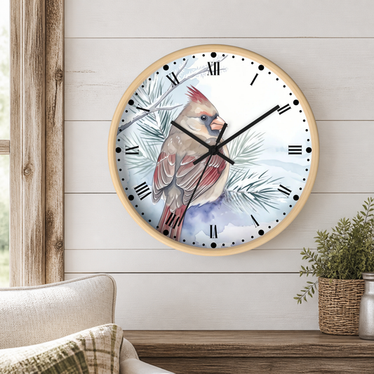 A round wall clock with a light wood frame hangs on a white shiplap wall above a wooden table. The clock face features a painted cardinal perched among pale blue-gray pine branches against a light blue and white background.