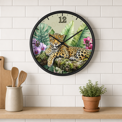 A round wall clock with a black frame hangs on a white subway tile wall above a wooden shelf. The clock face features a detailed illustration of a leopard surrounded by tropical green leaves and pink flowers.