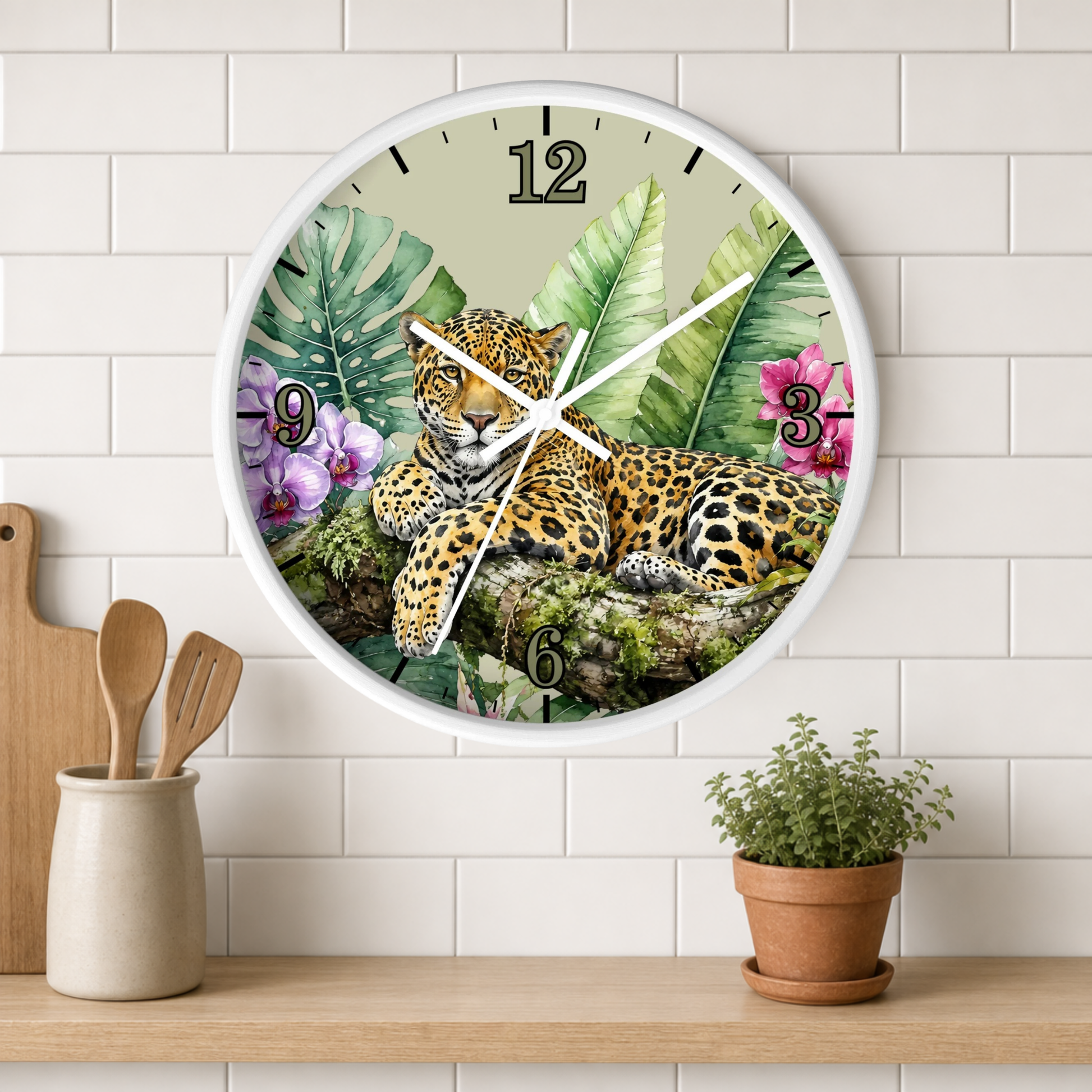 A round wall clock with a white frame hangs on a white subway-tile wall above a wooden shelf. The clock face features a detailed illustration of a leopard resting among tropical green leaves and pink flowers.