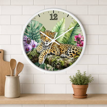 A round wall clock with a white frame hangs on a white subway-tile wall above a wooden shelf. The clock face features a detailed illustration of a leopard resting among tropical green leaves and pink flowers.