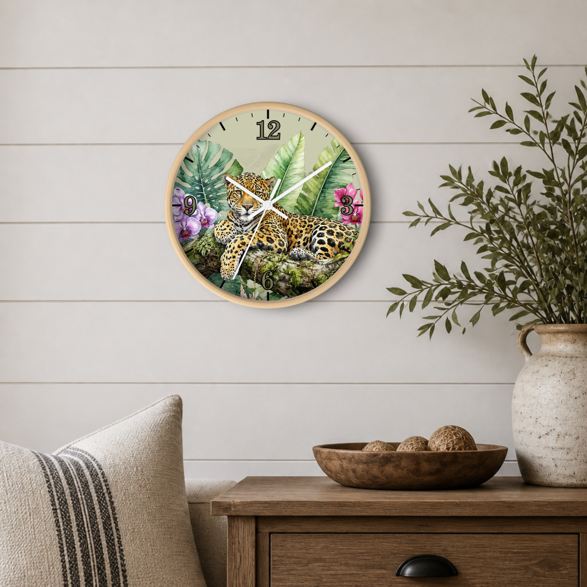 A round wall clock featuring a tropical jungle scene with a spotted big cat, displayed on a light wood-paneled wall above a wooden side table with a vase and bowl.